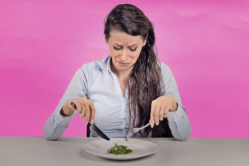 Female sitting at a table eating salad leaves with disgusted look on her face.