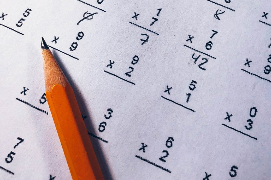 An orange pencil ontop of a sheet of paper with mathematic sums written